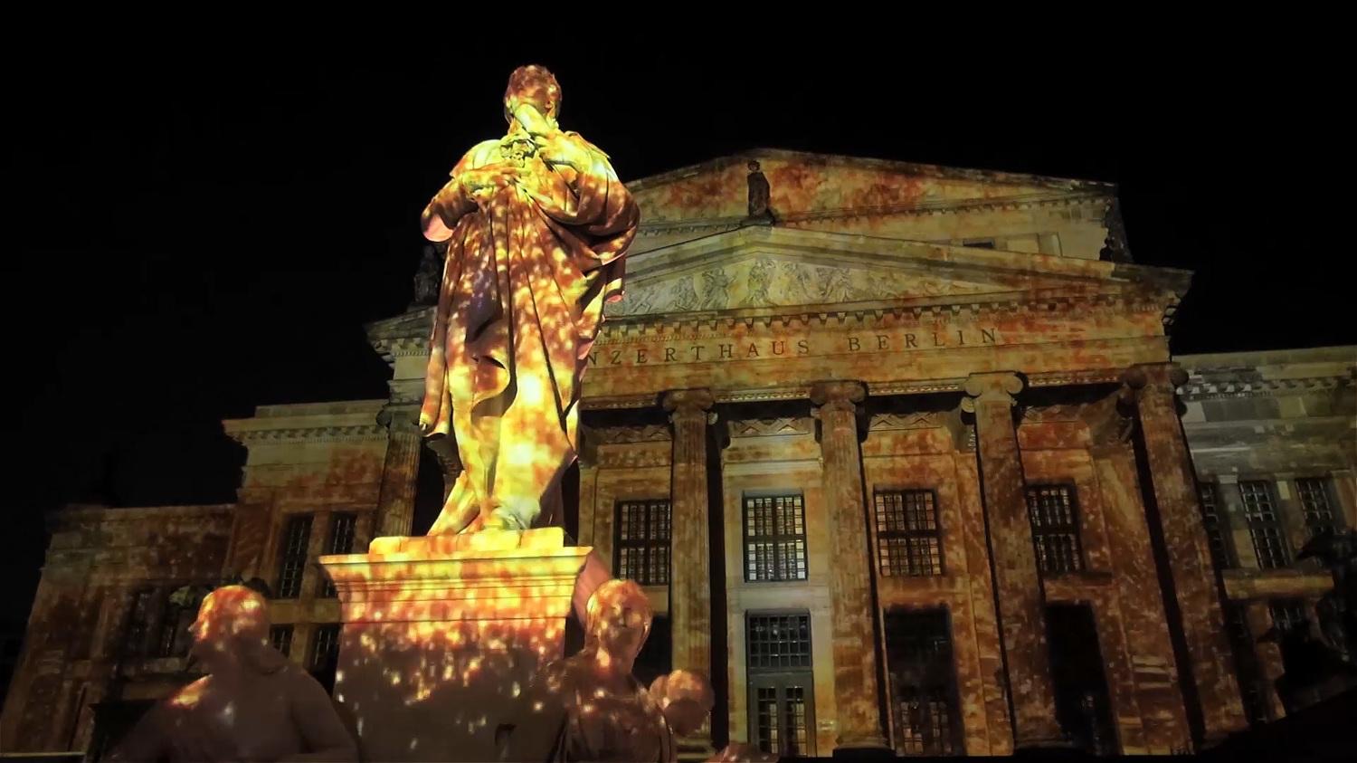The columned façade of the Konzerthaus at Gendarmenmarkt in Berlin lit by a colourful projection mapping with the Schiller monument in front