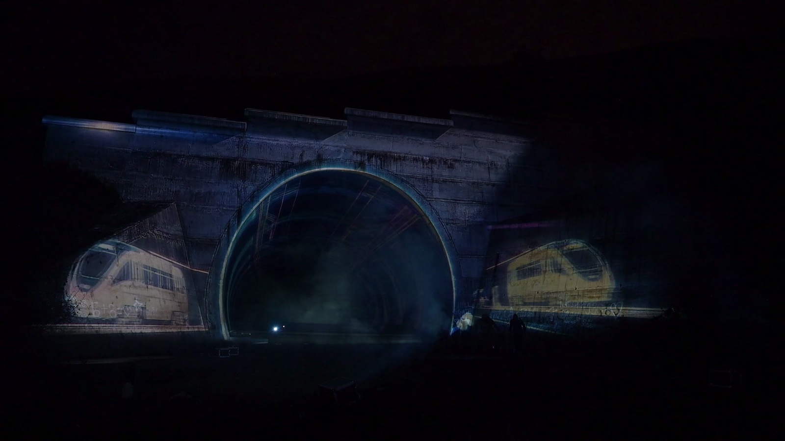 A stone railway tunnel portal in Tbilisi covered in a projection mapping of coloured tiles, with a hologram of light appearing in the tunnel mouth
